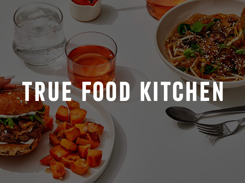 True Food Kitchen logo over an image of delicious food on white plates on a white table with a cocktail and glass of water.